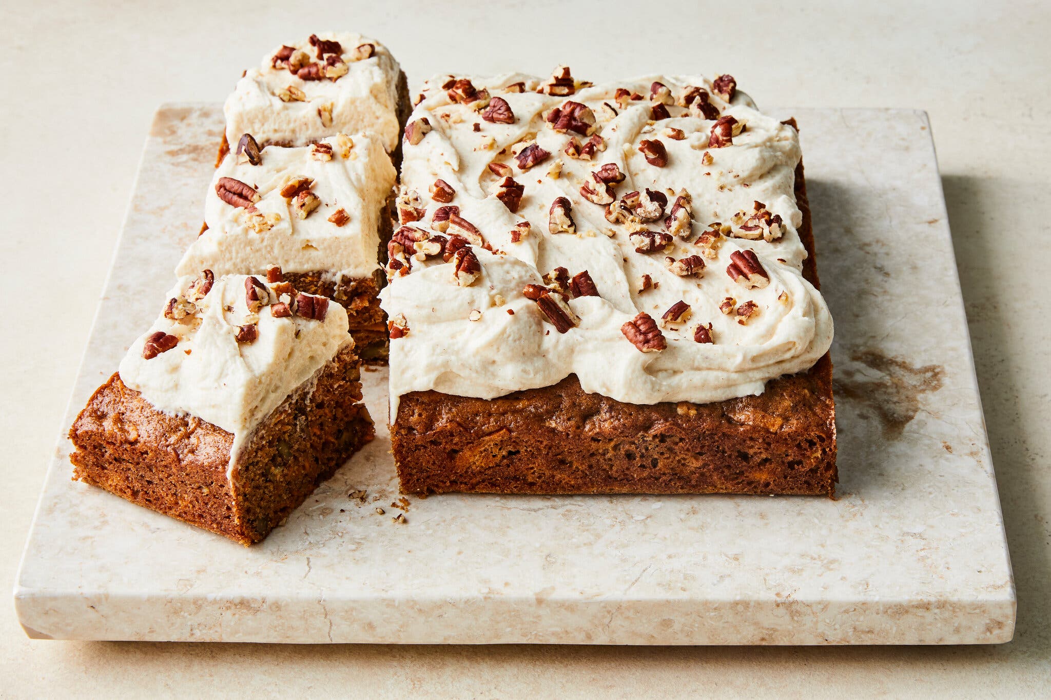 A slab of cake, topped with a pillowy frosting and pecans is photographed at an angle. Three square slices sit off to the side.