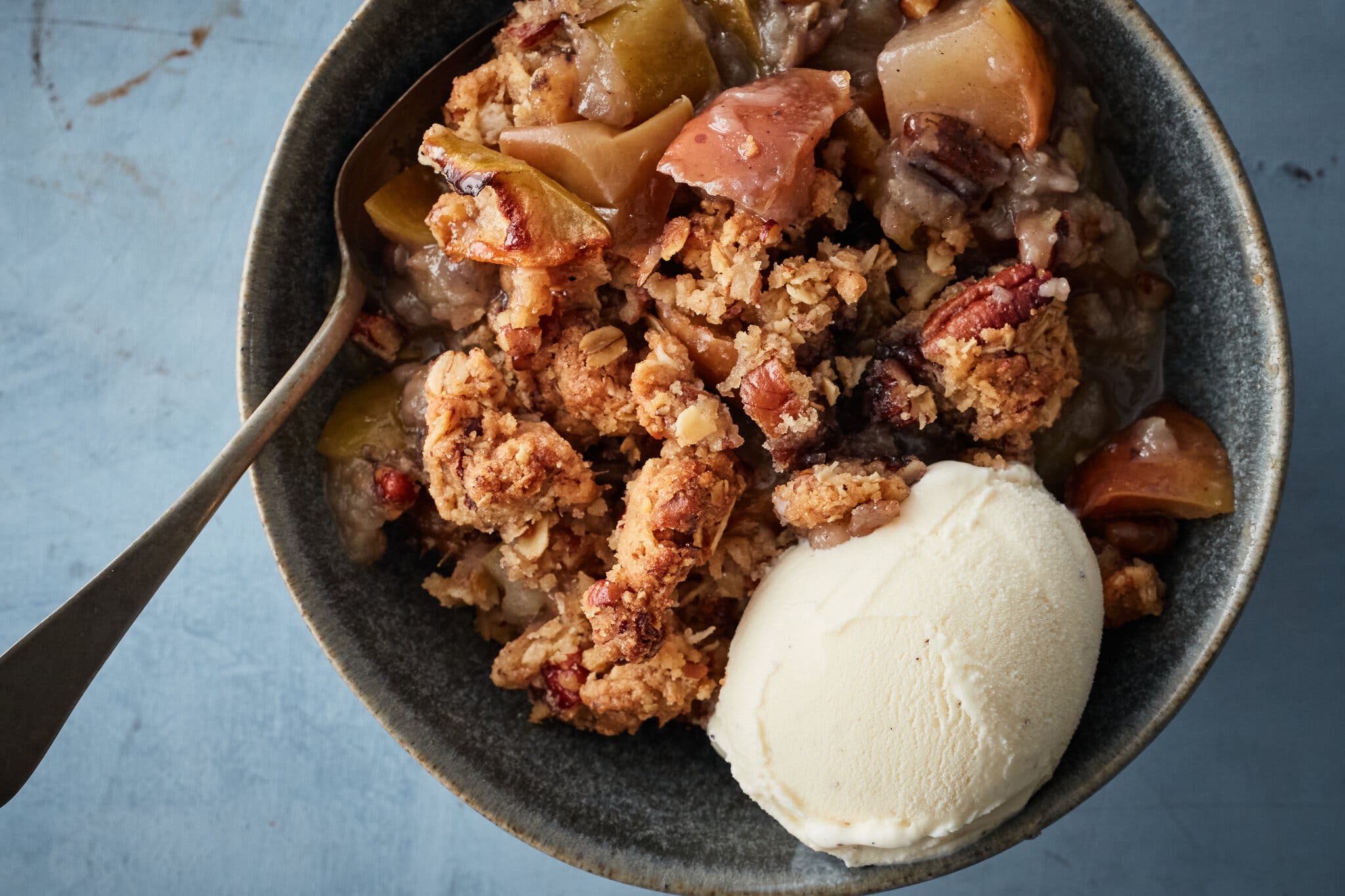 An overhead shot of a dark bowl of apple crisp with a scoop of ice cream. A metal spoon juts out at the top left corner.