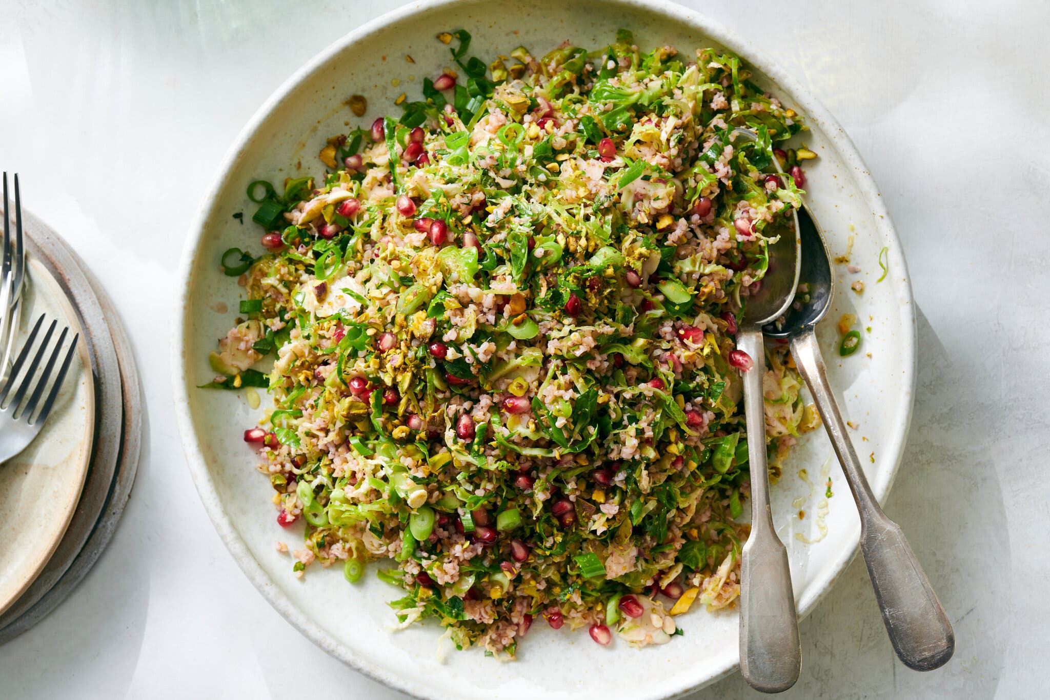 An overhead shot of a white plate of brussels sprout salad topped with pomegranate seeds and pistachios. Utensils sit on the right side of the plate.