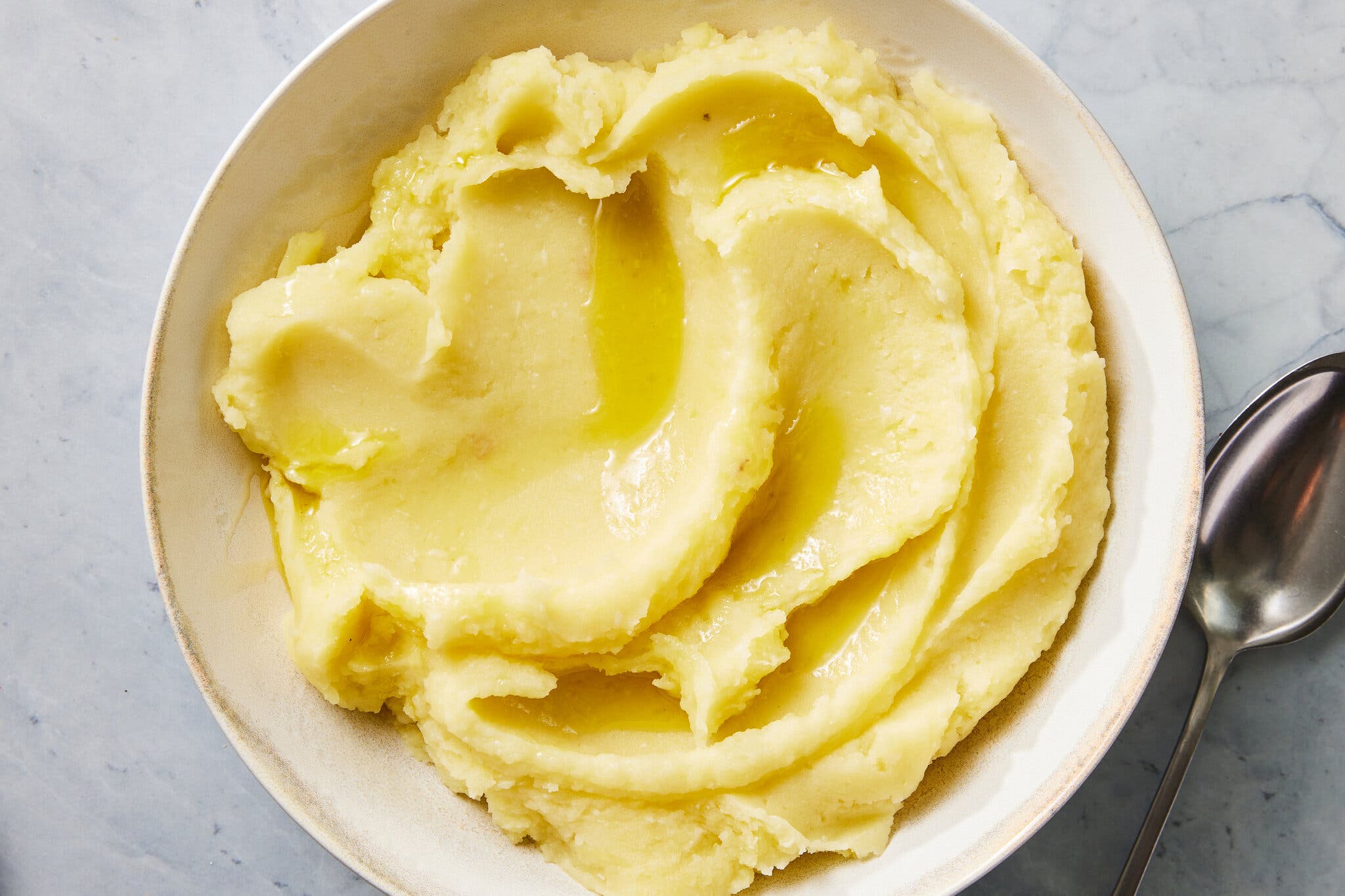 An overhead shot of a bowl of mashed potatoes topped with olive oil. A metal spoon sits next to the bowl.