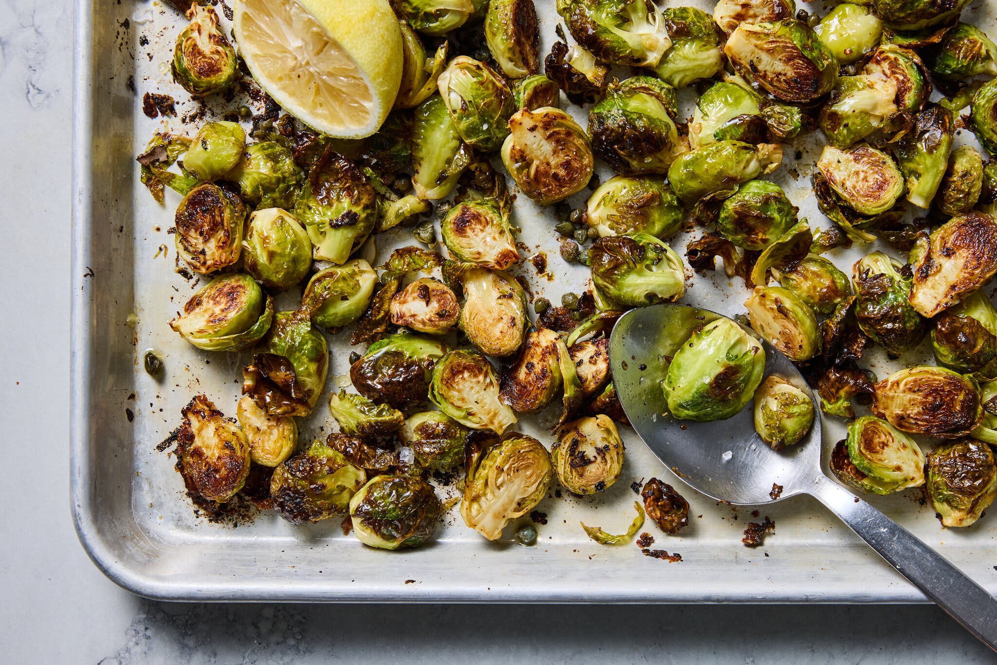A sheet pan of roasted brussels sprouts. A metal spoon sits at the bottom right corner.